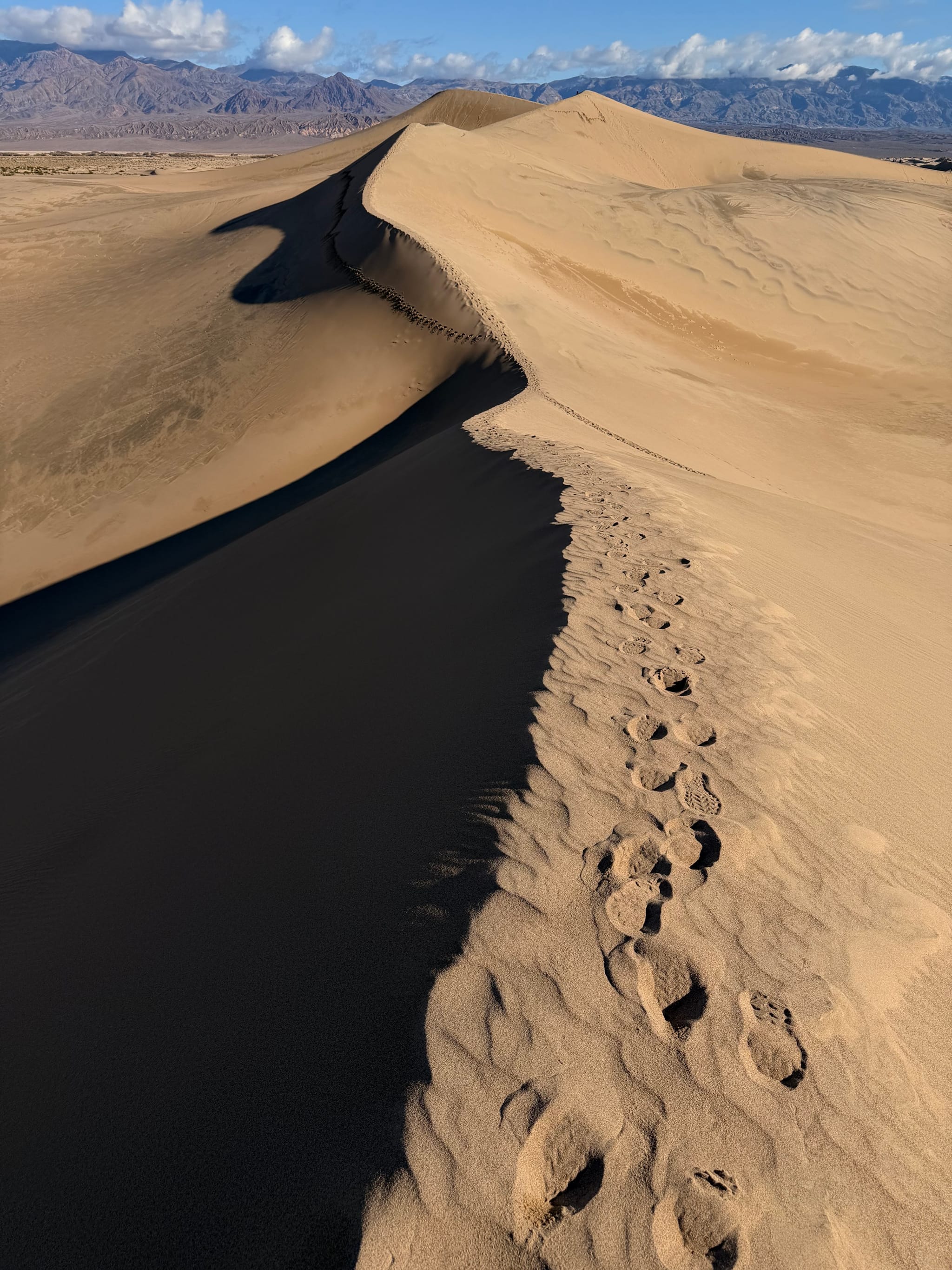 Sand dune crest with a line of footprints along the ridge, casting a long shadow across the slope with distant mountains on the horizon