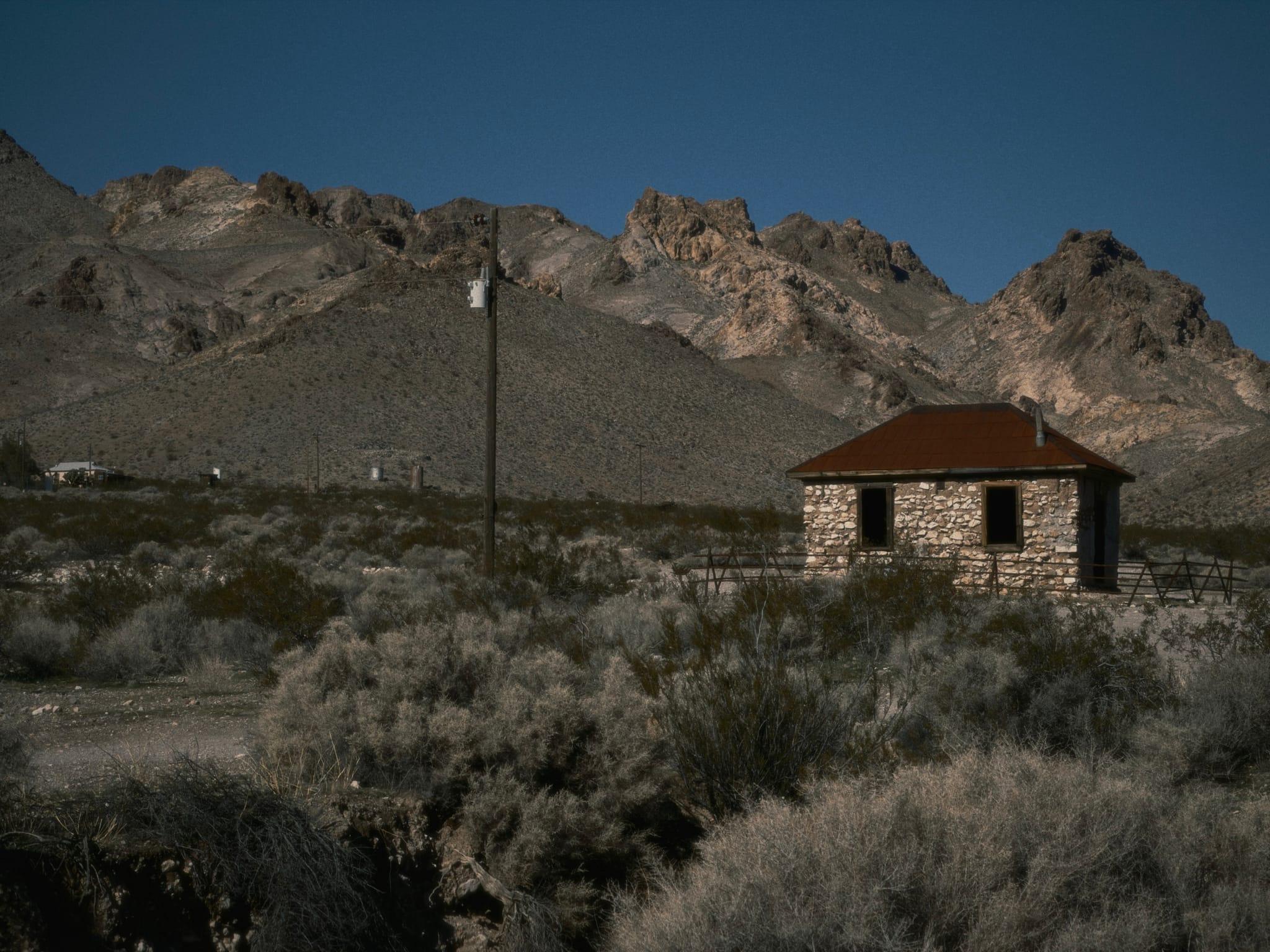 A small stone cabin sits in a scrubby desert foreground with a utility pole nearby and rugged rocky mountains in the background under a clear blue sky