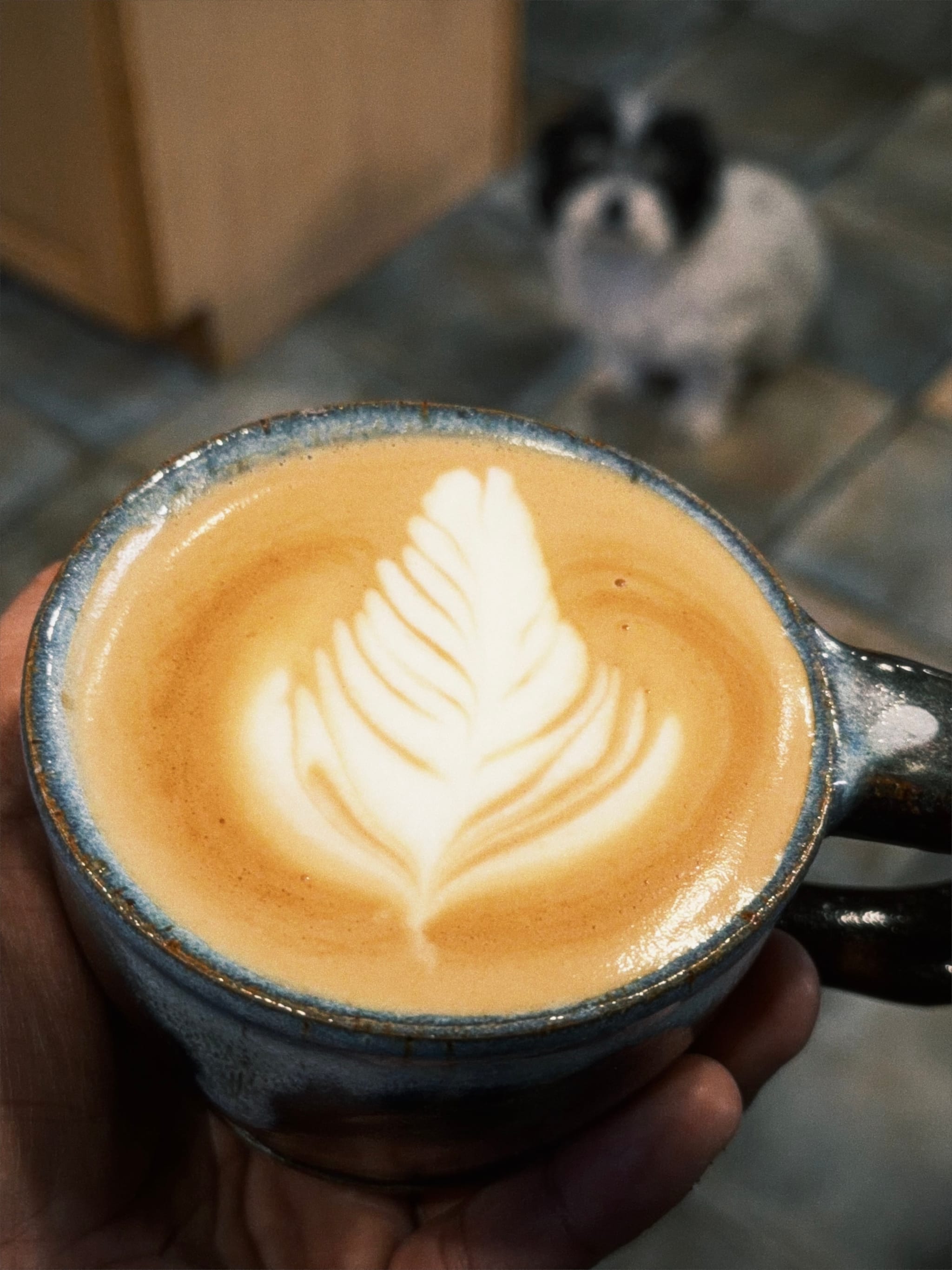 Hand holding a ceramic cup of latte with leaf-shaped foam art while a small dog sits blurred in the background on a tiled floor