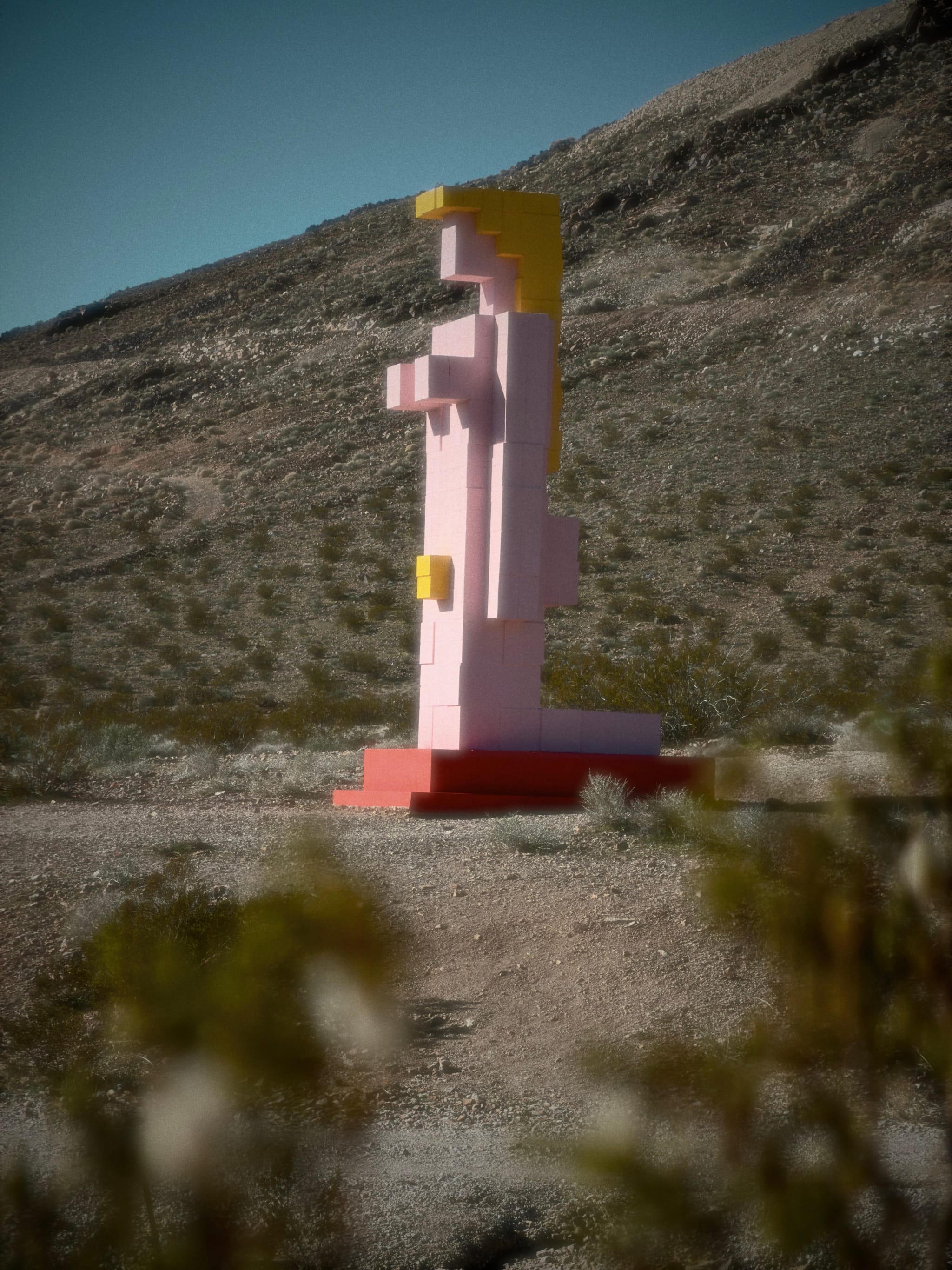 A tall pink block-style sculpture on a red base stands in a rocky desert landscape with a hillside backdrop