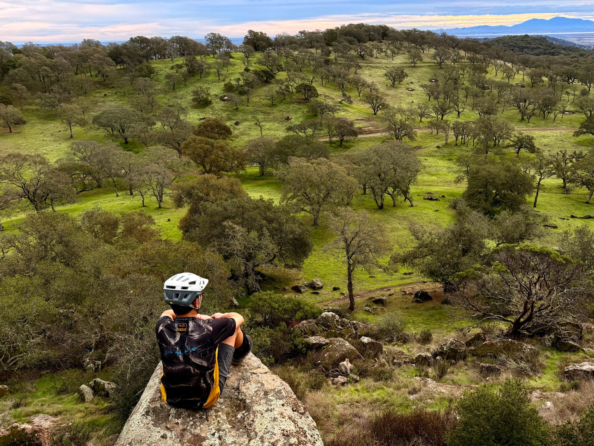 Cyclist with a backpack and helmet sits on a large rock overlooking rolling green hills dotted with trees under a cloudy sky